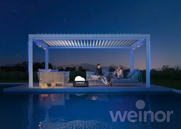 Beleuchtete Pergola am Pool, Paar sitzt auf Loungemöbeln unter Lamellendach.